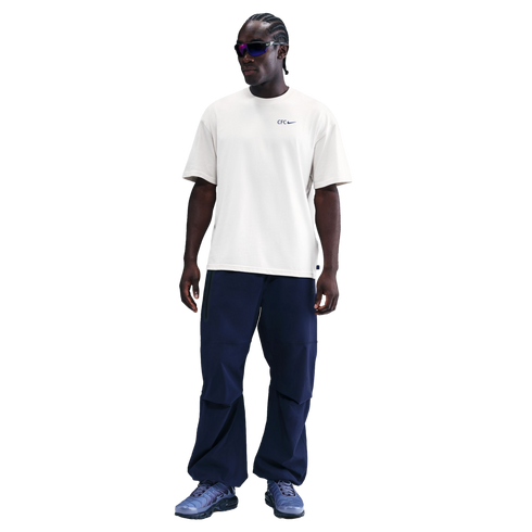 A man wearing a cream Nike CFC t-shirt, navy cargo pants, and blue sneakers against a black background.