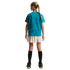 Back view of a girl with a blonde braid wearing a teal patterned t-shirt, beige pleated skirt, black knee-high socks, and black sneakers against a black background.
