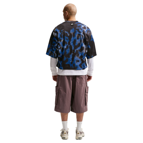 Rear view of a man wearing a blue and black patterned mesh tee layered over a white long-sleeve shirt, mauve cargo shorts, and white sneakers.