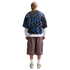 Rear view of a man wearing a blue and black patterned mesh tee layered over a white long-sleeve shirt, mauve cargo shorts, and white sneakers.