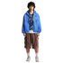 A man with curly hair poses in a blue windbreaker, a patterned top, brown cargo shorts, and sneakers against a plain white background.