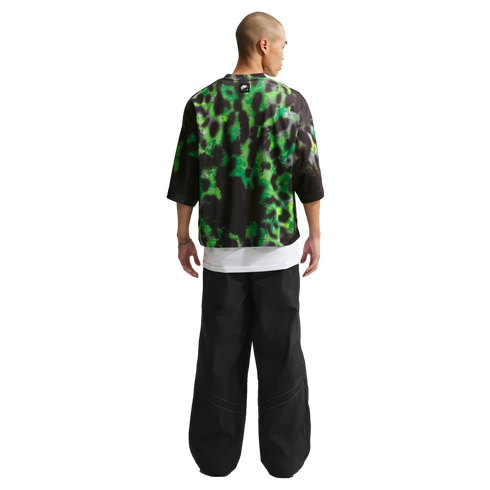 Back view of a person wearing a green and black mesh top over a white tee and baggy black trousers against a white backdrop.