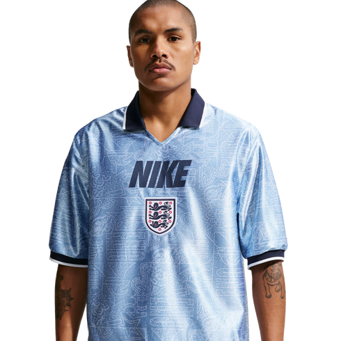A man wearing a light blue, patterned Nike England football jersey with a navy blue collar and the Three Lions crest, posed against a white background.