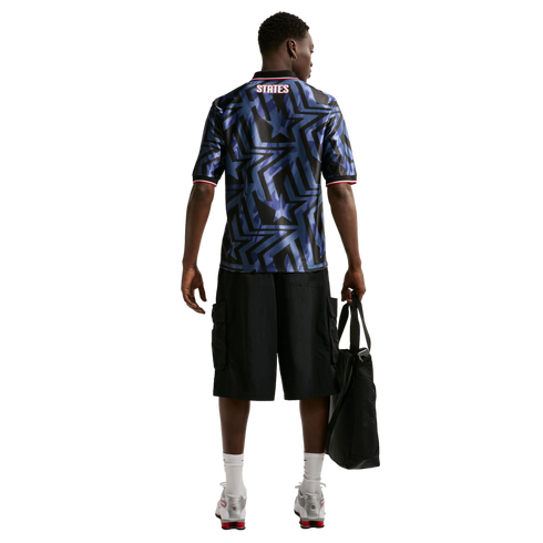 Rear view of a man wearing a blue star-patterned jersey with "STATES" on the back, black shorts, and silver sneakers, holding a black bag.