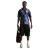 A Black man wearing a blue patterned USA soccer jersey, black cargo shorts, and sneakers stands holding a black bag against a white background.