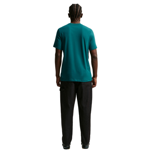 A rear view of a man with braided hair wearing a teal t-shirt and dark, wide-leg pants against a solid black background.