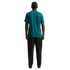 A rear view of a man with braided hair wearing a teal t-shirt and dark, wide-leg pants against a solid black background.