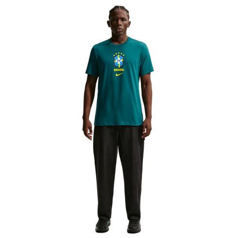 A young man stands centered against a solid black background, wearing a teal green Nike t-shirt featuring the Brazilian football federation (CBF) crest. He is also wearing dark charcoal straight-leg trousers and black sneakers.