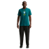 A young man stands centered against a solid black background, wearing a teal green Nike t-shirt featuring the Brazilian football federation (CBF) crest. He is also wearing dark charcoal straight-leg trousers and black sneakers.