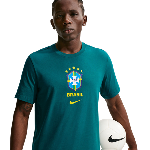 A young man wearing a teal Nike t-shirt with the Brazil national soccer team crest, holding a white soccer ball against a solid black background.