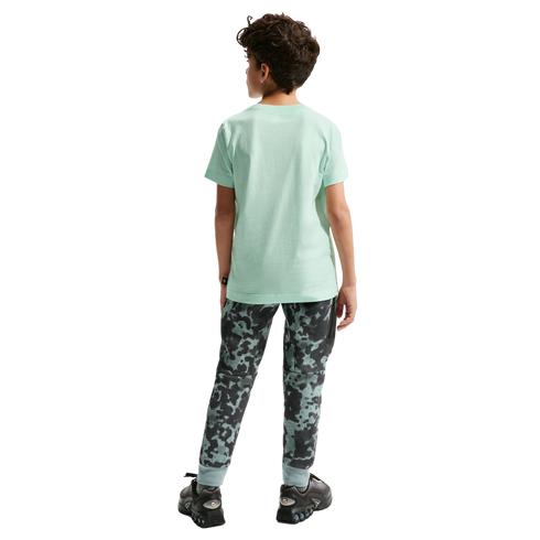 Back view of a young boy with curly brown hair standing against a black background. He is wearing a light green short-sleeved t-shirt, camouflage-patterned joggers, and black athletic shoes.