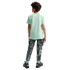 Back view of a young boy with curly brown hair standing against a black background. He is wearing a light green short-sleeved t-shirt, camouflage-patterned joggers, and black athletic shoes.