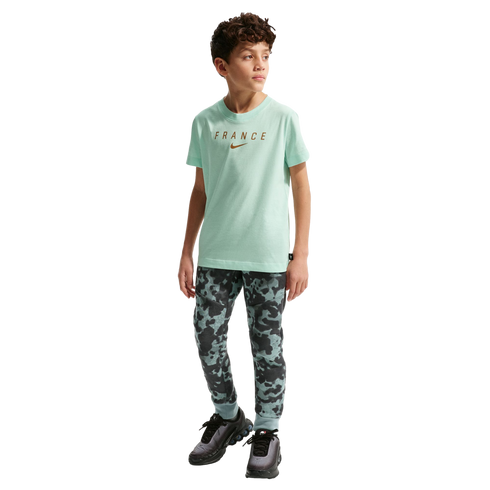 A young boy stands against a solid black background. He is wearing a light mint green short-sleeved T-shirt with the word "FRANCE" and a Nike swoosh in gold, patterned camouflage joggers, and black sneakers.