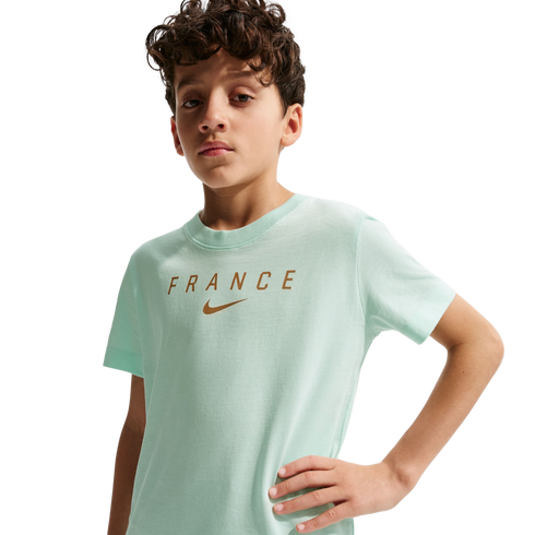 A young boy with curly brown hair wearing a light mint green t-shirt with "FRANCE" and a Nike swoosh printed in gold across the chest. He stands with one hand on his hip against a solid black background.