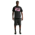 A man stands against a white background wearing a black Nike "Friendship Tournament" graphic t-shirt, charcoal shorts, and black sneakers.