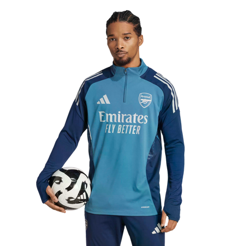 A man with braided hair wears a teal Arsenal training top and navy pants, holding a soccer ball under his arm against a black background.