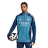 A man with braided hair wears a teal Arsenal training top and navy pants, holding a soccer ball under his arm against a black background.