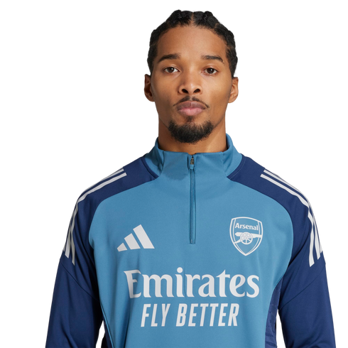 Headshot of a man with braided hair and a goatee wearing a blue Arsenal training zip-up top with the Emirates sponsor logo, set against a solid black background.