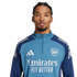 Headshot of a man with braided hair and a goatee wearing a blue Arsenal training zip-up top with the Emirates sponsor logo, set against a solid black background.