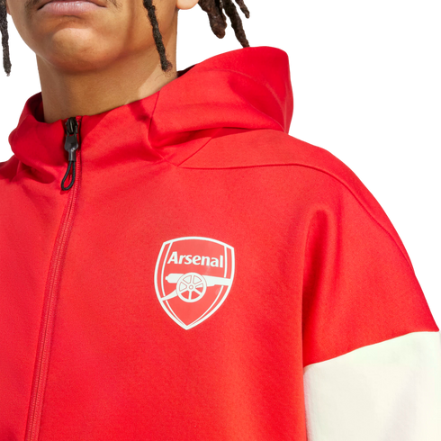 A person with dreadlocks wearing a red zip-up hoodie featuring the Arsenal football club crest on the chest and white color-block sleeves.