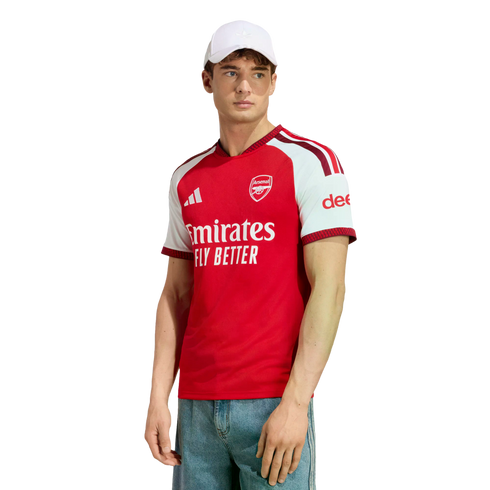 A man poses in a red and white Arsenal football jersey, light blue jeans, and a white baseball cap against a white background.