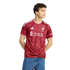 A man with curly hair wearing a red and white Liverpool FC soccer jersey and blue jeans, standing against a white background.
