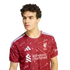 Man with curly brown hair wearing a red Liverpool FC soccer jersey, looking to the side against a plain white background.