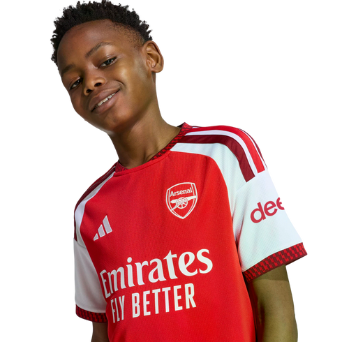 A smiling young Black boy wearing a red and white Arsenal football jersey against a white background.