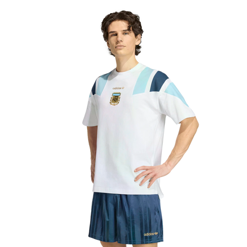 A man wears a white Adidas Argentina t-shirt and blue shorts, posing against a white background.
