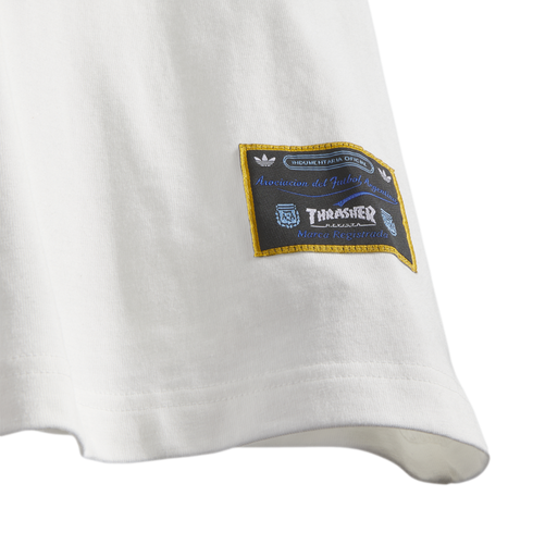 Close-up of a white shirt hem featuring a black and gold woven patch with logos for Adidas, Thrasher magazine, and the Argentine Football Association (AFA).