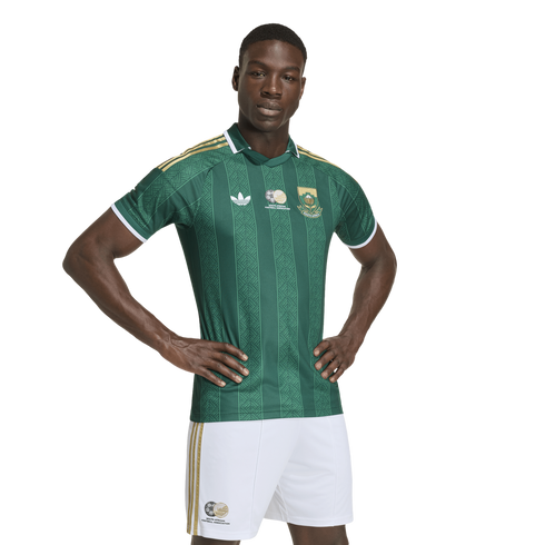 A Black man models a green and gold South African national team soccer kit, featuring a patterned jersey and white shorts, against a white background.