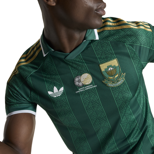 A person wearing a green South Africa national football jersey with gold shoulder stripes, a white Adidas logo, and the national team crest over a patterned fabric.