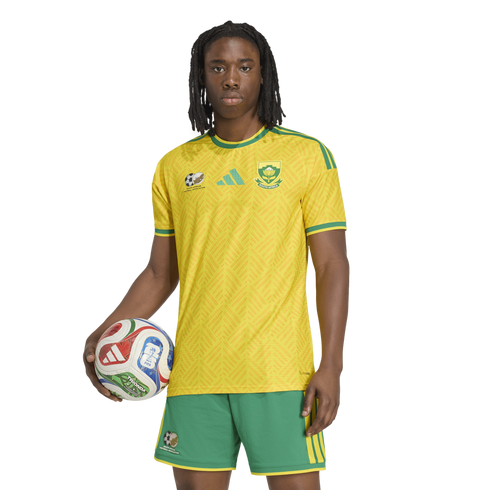 A person with dreadlocks wearing a yellow South African soccer kit, holding a soccer ball against a white background.