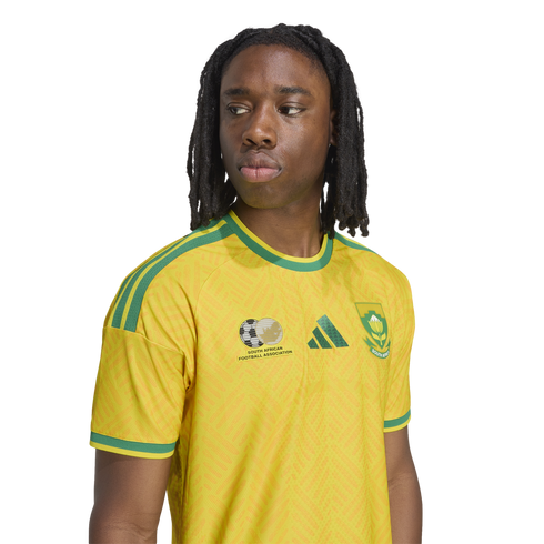 A person with long dreadlocks wearing a yellow and green South African national soccer jersey, looking to the side against a white background.
