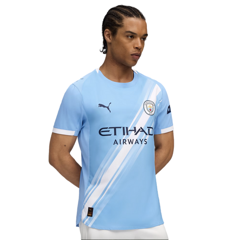 A man models a light blue Manchester City soccer jersey featuring a white diagonal sash, team crest, and sponsor logos.