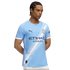 A man models a light blue Manchester City soccer jersey featuring a white diagonal sash, team crest, and sponsor logos.