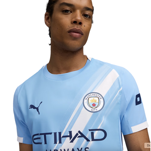 A person wearing a light blue Manchester City football jersey with a white diagonal sash, the club crest, and a Puma logo.