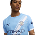A person wearing a light blue Manchester City football jersey with a white diagonal sash, the club crest, and a Puma logo.