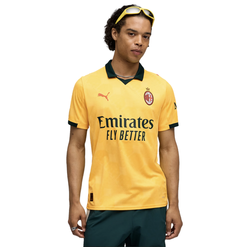 A man wearing a yellow AC Milan soccer jersey, dark green shorts, and yellow sunglasses on his head, posed against a black background.