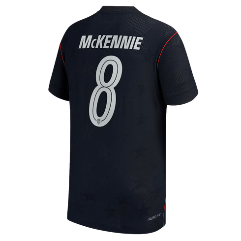 The back view of a navy blue soccer jersey featuring the name "MCKENNIE" and the number "8" in large white lettering. The jersey has a subtle tonal star pattern across the fabric and thin red piping along the shoulder and side seams.