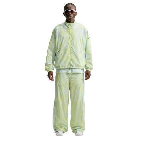 A person wearing a light green and white patterned Nike tracksuit, futuristic white sunglasses, and white sneakers against a white background.