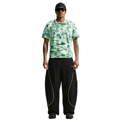 Man wearing a green patterned Nike shirt, baggy black pants with lime yellow piping, a black beanie, and sunglasses against a plain white background.