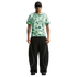 Man wearing a green patterned Nike shirt, baggy black pants with lime yellow piping, a black beanie, and sunglasses against a plain white background.