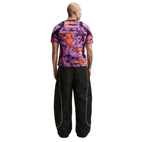Rear view of a man in a vibrant purple and orange patterned shirt and oversized black track pants with neon green piping, against a plain white background.