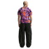 Rear view of a man in a vibrant purple and orange patterned shirt and oversized black track pants with neon green piping, against a plain white background.