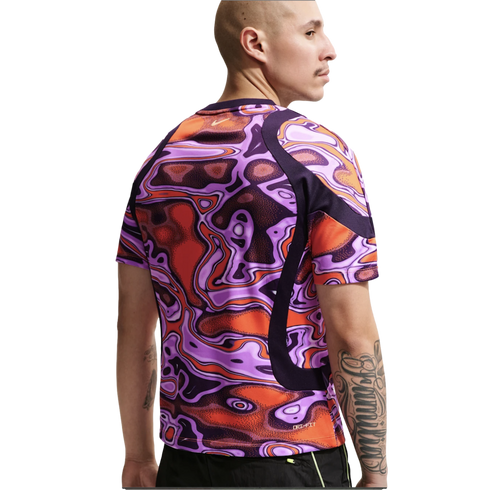 Rear view of a man wearing a Nike shirt with a vibrant, abstract purple and orange pattern. He has a bald head and arm tattoos, set against a plain white background.